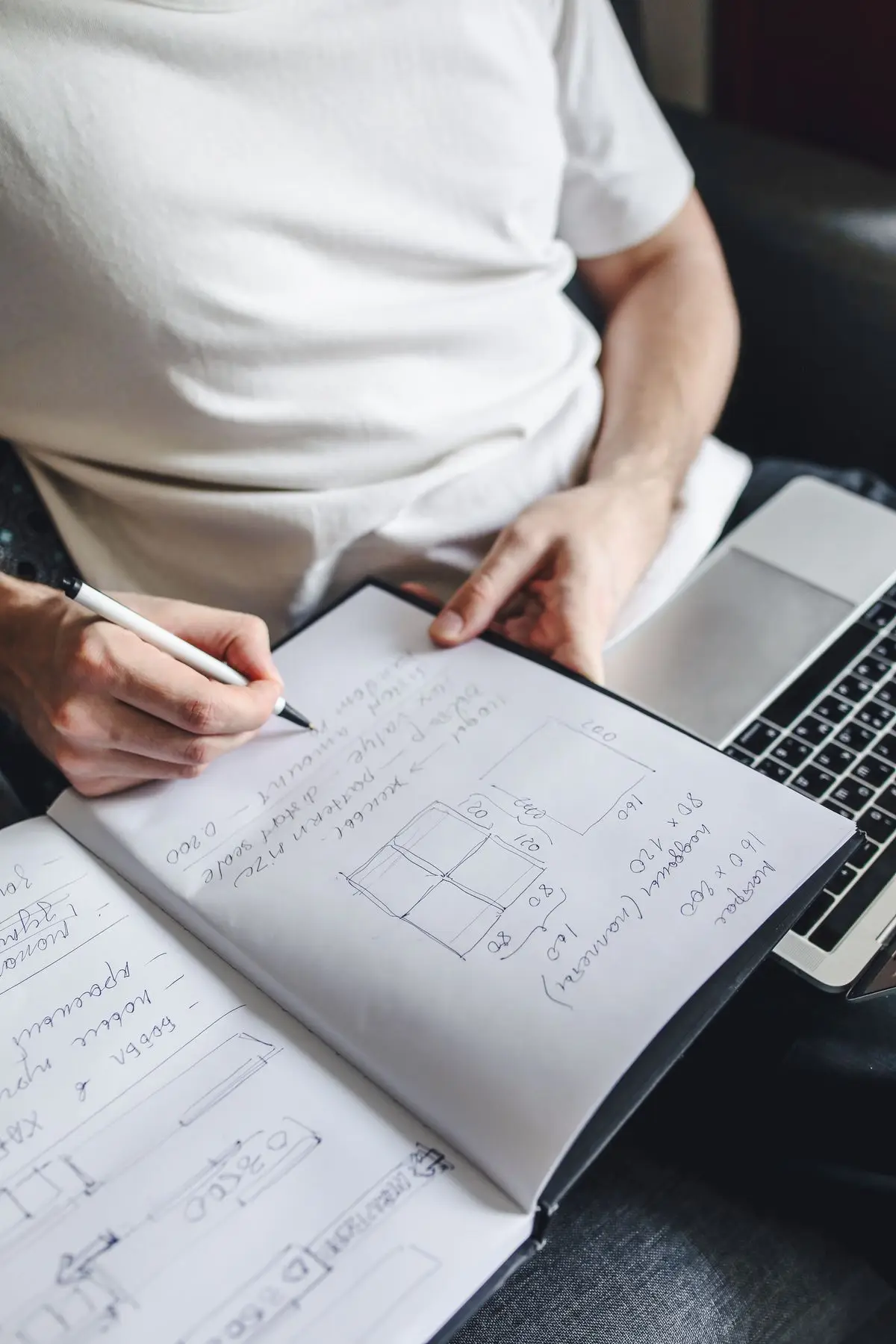 Hands writing in a notebook next to a laptop on a couch.