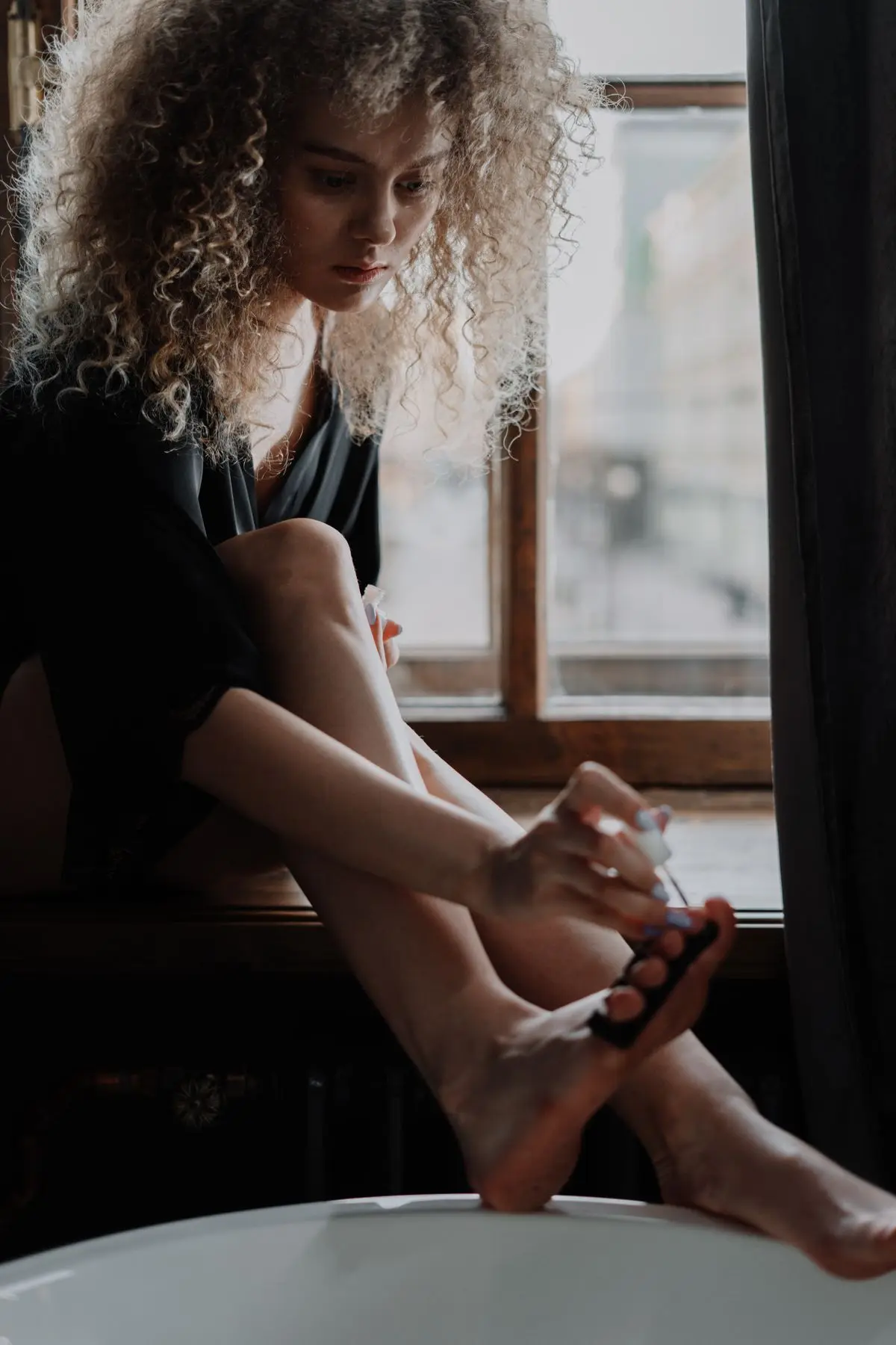 A young woman painting her toenails while sitting by a window.
